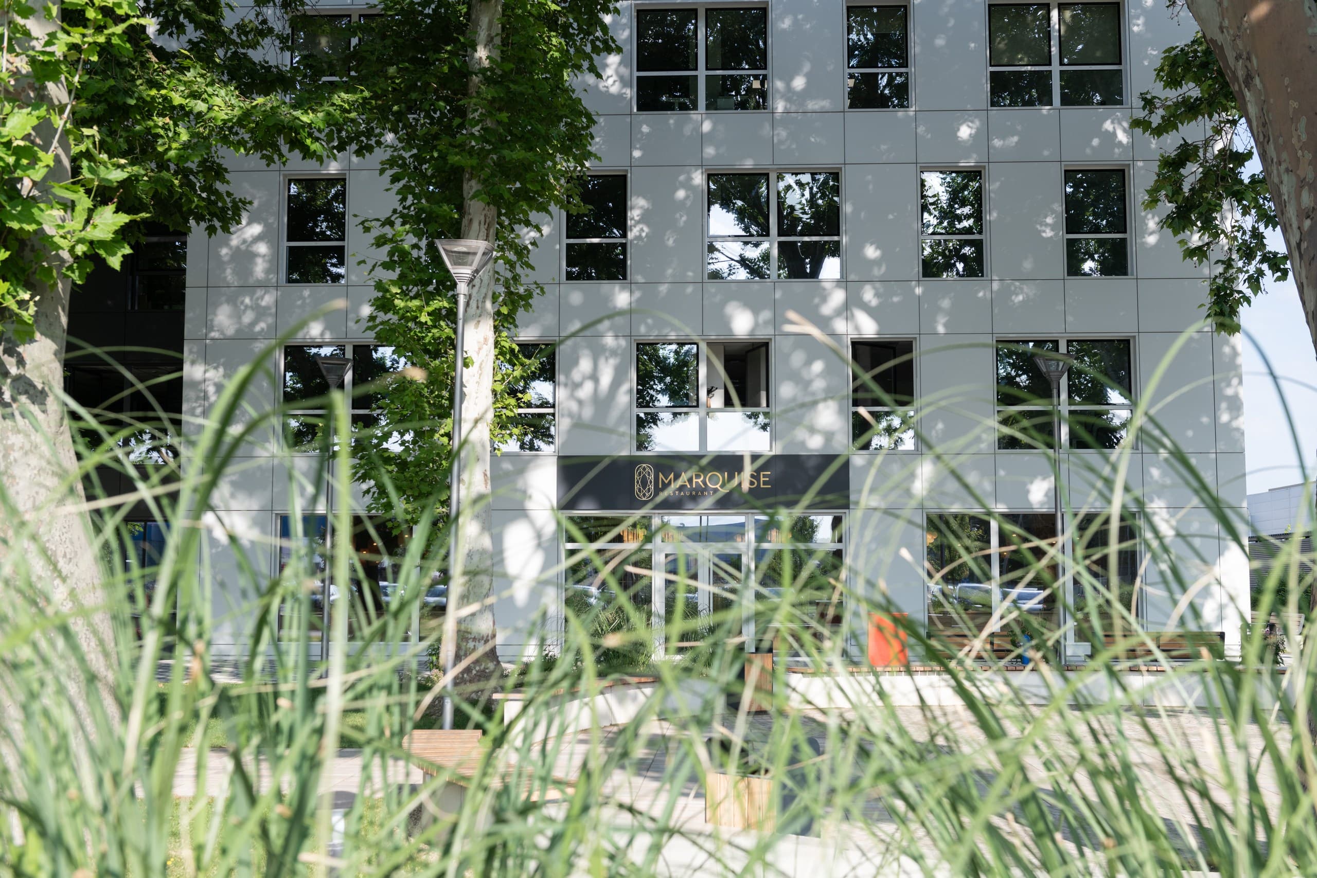 Entrance to Marquise restaurant on a modern white building, seen through tall green grass.