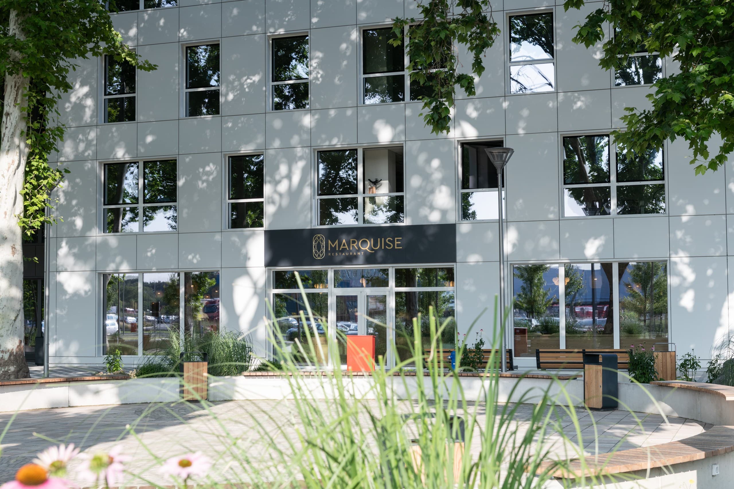 Modern white building facade featuring Marquise Restaurant with a paved courtyard and lush greenery.