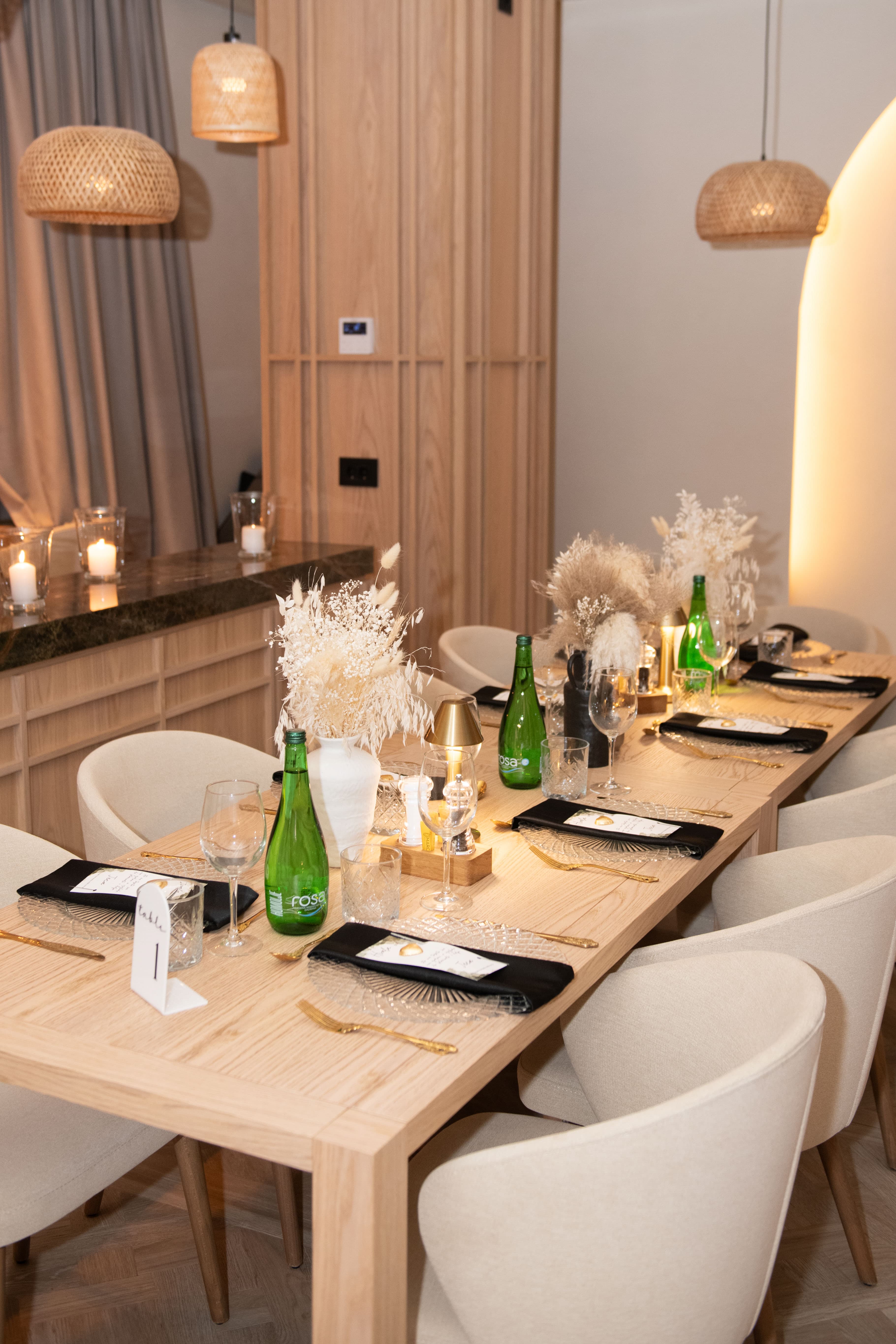 Elegant wooden dining table set with black napkins, gold cutlery, and dried floral centerpieces.
