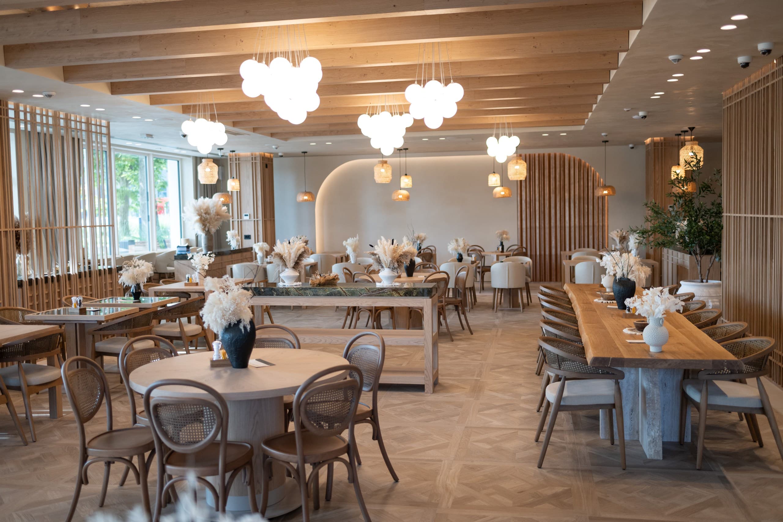 Modern restaurant interior featuring light wood furniture, cloud-shaped pendant lights, and dried floral arrangements.