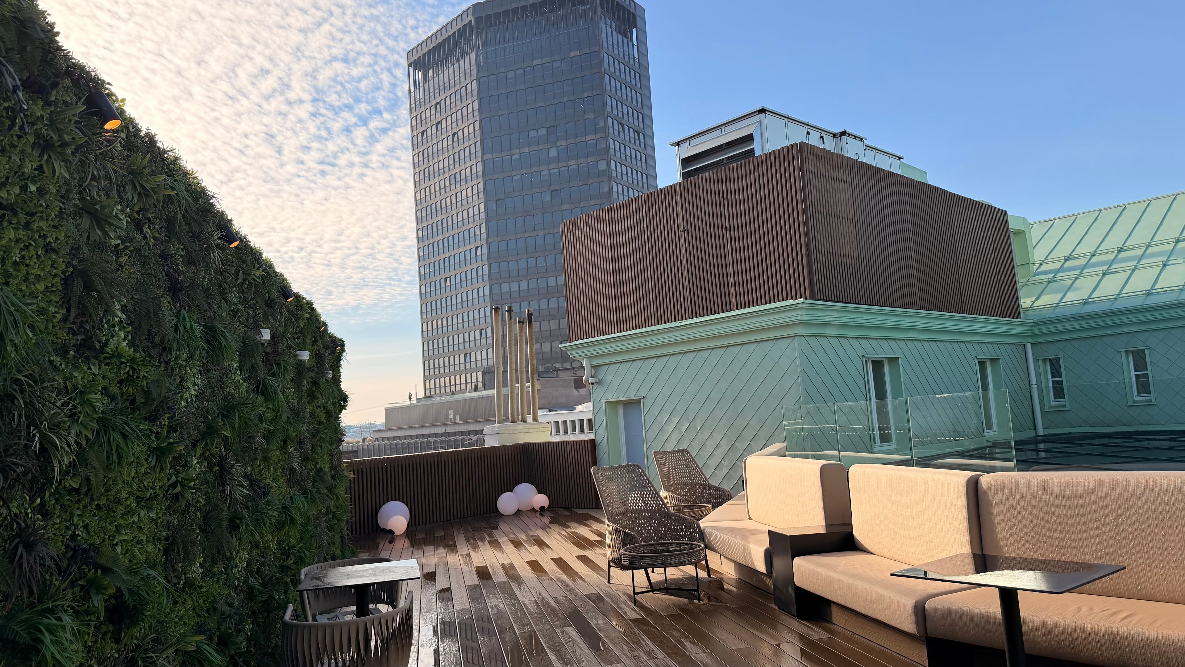 Modern rooftop lounge featuring wooden decking, a vertical garden, and a tall city skyscraper.