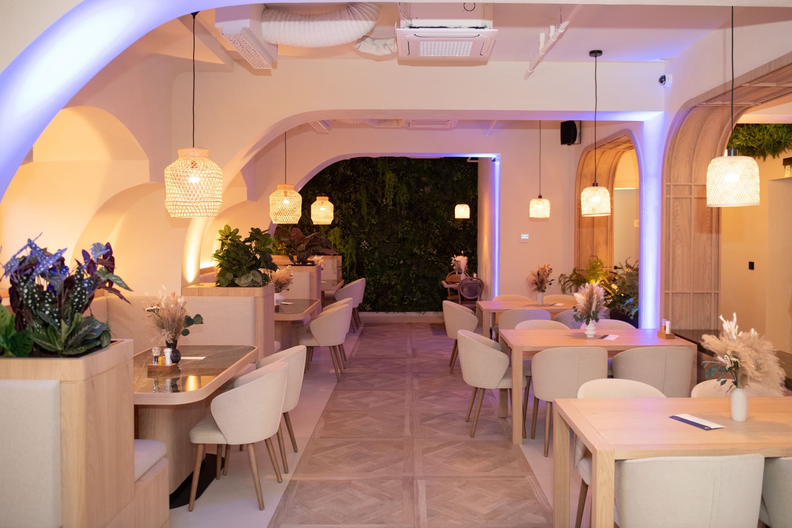 Modern restaurant interior with arched walls, woven pendant lights, wooden tables, and lush green plants.