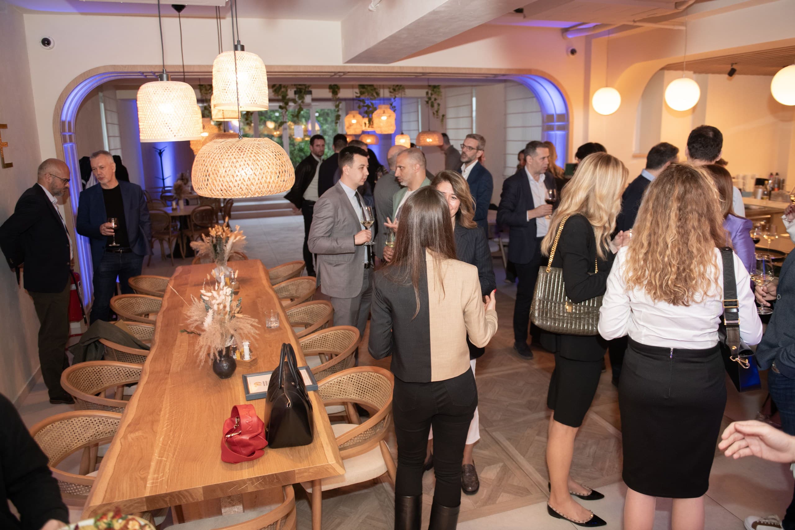 Professionals socialize in a modern restaurant featuring long wooden tables and warm woven pendant lights.