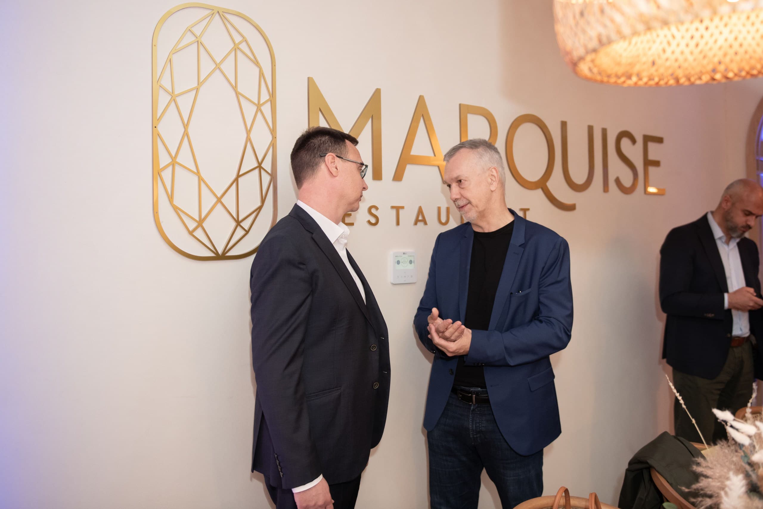 Two men in professional attire converse in front of a gold Marquise Restaurant wall logo.